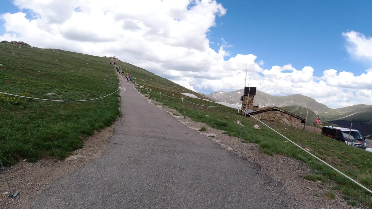 A paved trail in Winter Park, Colorado climbs a grassy slope toward distant hikers, with rope railings, parked cars, a small structure, and cloud-filled sky visible.