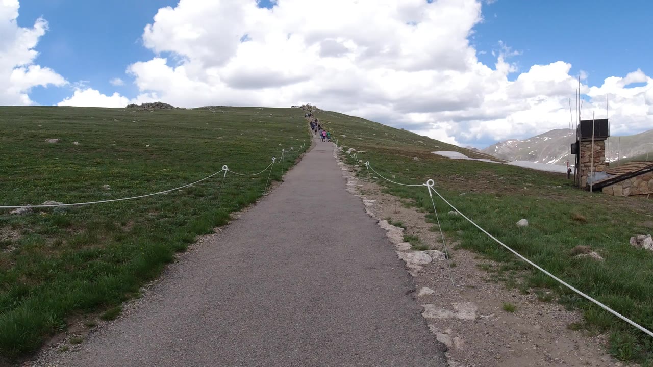 Paved mountain path through grassy hills under bright clouds.