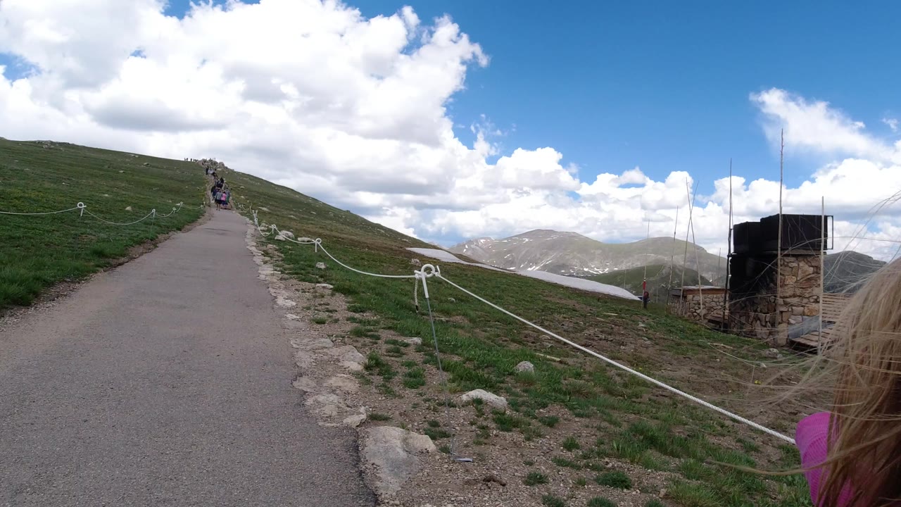A Winter Park, Colorado uphill path runs between grassy slopes and rope fencing, with a child's hair and pink sleeve partly visible at the right edge.