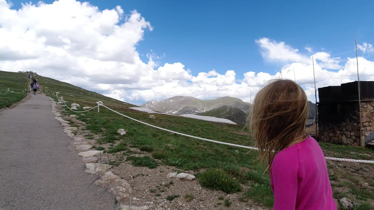 The foreground person in pink remains near the right edge while the paved Winter Park mountain path leads toward small hikers and cloud-covered peaks.