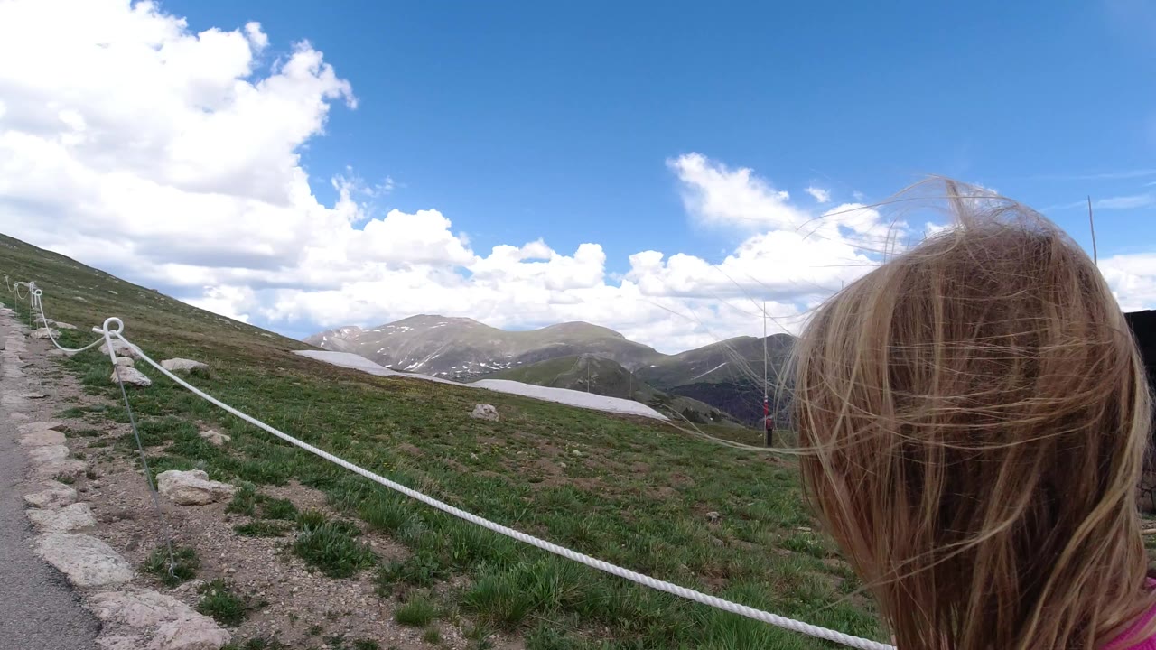 A blond child in a pink top fills the right foreground beside a roped gravel trail in Winter Park, Colorado, with green slopes, snow patches, distant peaks, and bright clouds beyond.