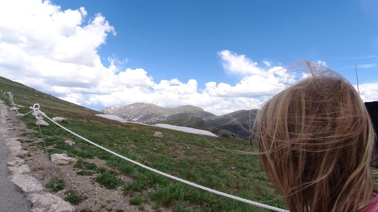 The child's windblown blond hair occupies the right foreground as a white rope barrier follows the Winter Park mountain path toward grassy slopes and distant peaks under a cloud-filled sky.
