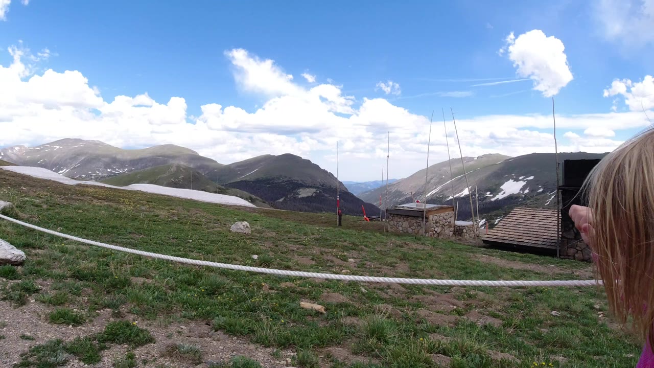 Mountain path area with rope fence, grass, snow patches, and a child partly visible at right.