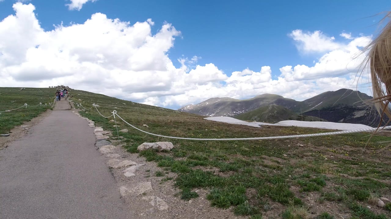 Paved mountain trail with hikers ahead, rope barrier, grass, mountains, and clouds.