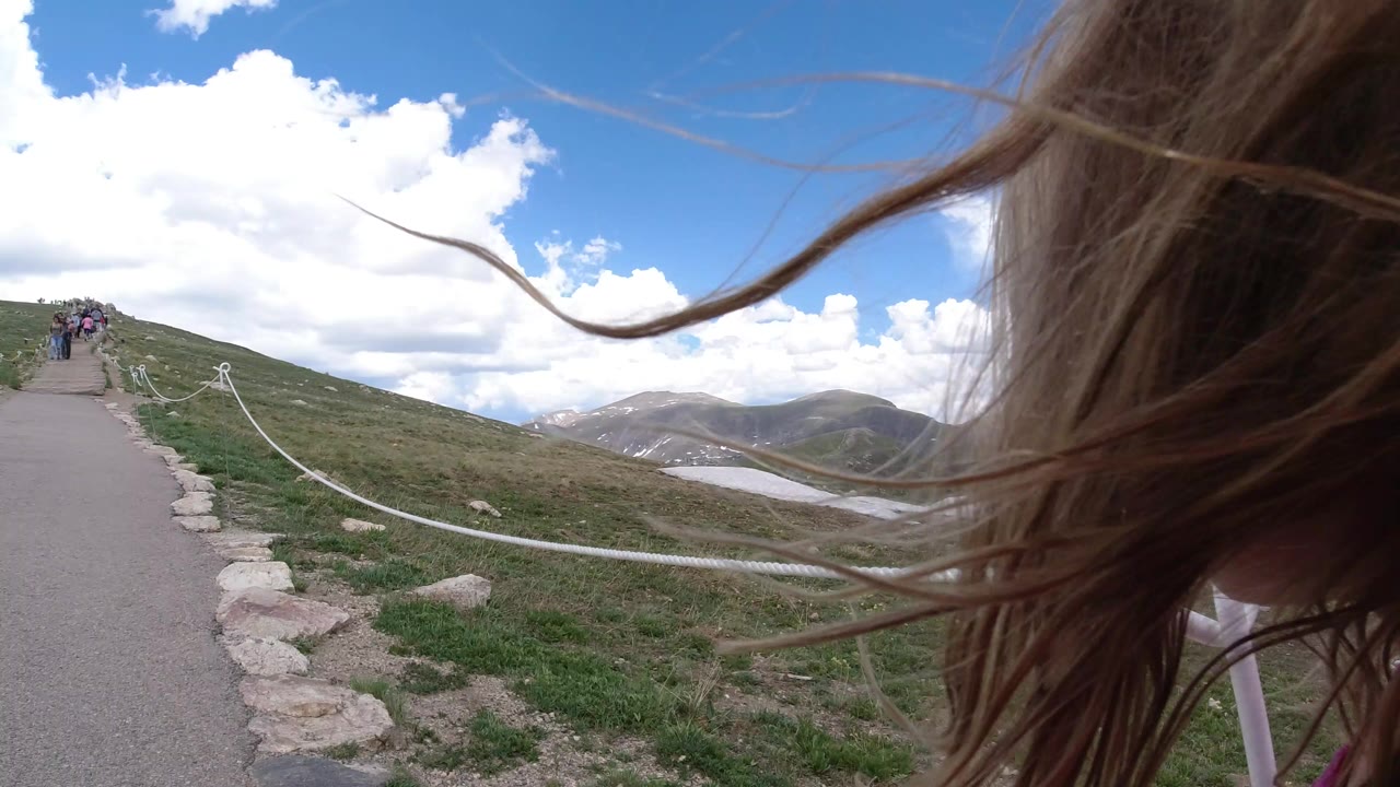A child’s windblown hair fills the right foreground in Winter Park, Colorado, while a paved path, rope fence, grassy slope, snow patches, distant mountains, and small figures ahead are visible.