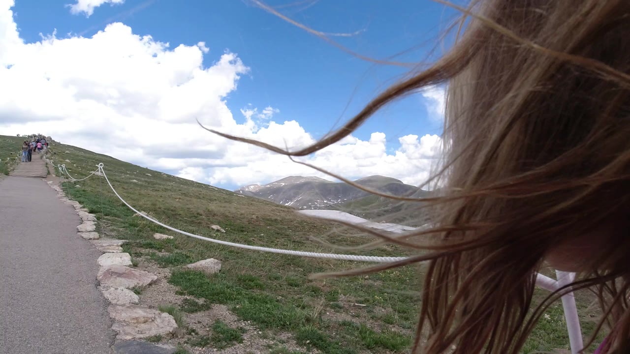 The child remains close at the right edge in Winter Park, Colorado, with hair sweeping across the frame above the path, rope railing, grass, snow patches, mountains, and bright clouds.