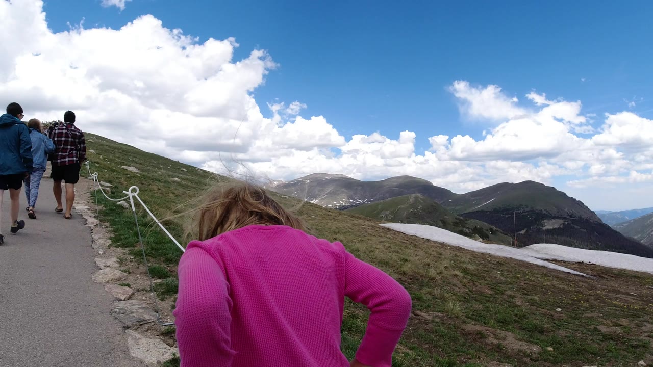 The child remains low in the foreground as several people walk ahead on the Winter Park mountain path, with a rope boundary and rolling ridges under bright clouds.