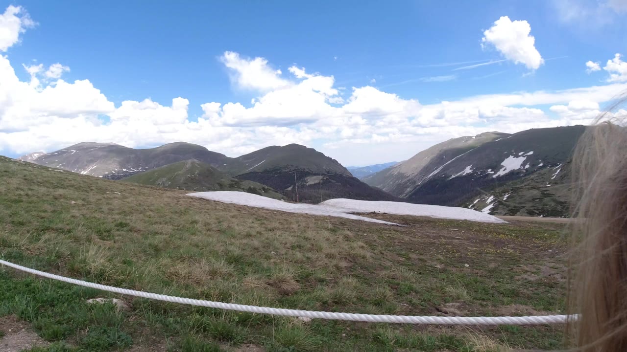 Mountain trail view with rope rail, grassy slopes, snow patches, and windblown hair near the lens.
