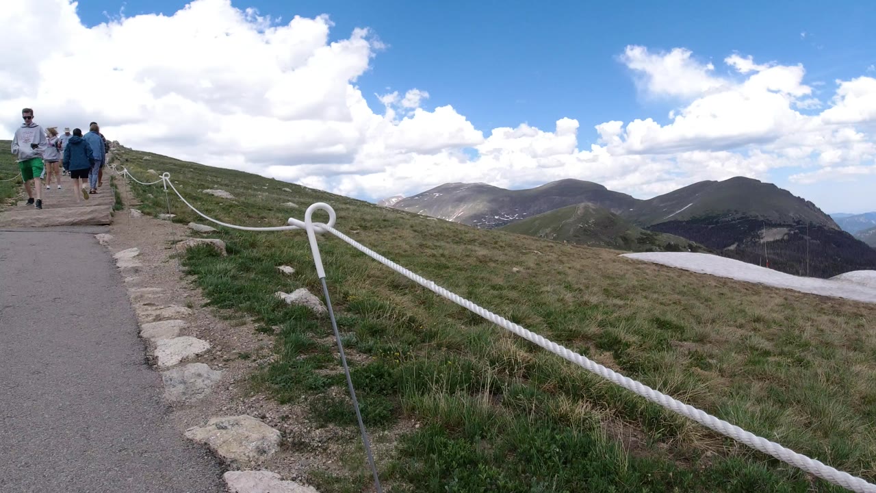 Hikers walk along a paved mountain path in Winter Park, Colorado, with white rope railing, grassy slopes, distant ridges, snow patches, and bright clouds overhead.