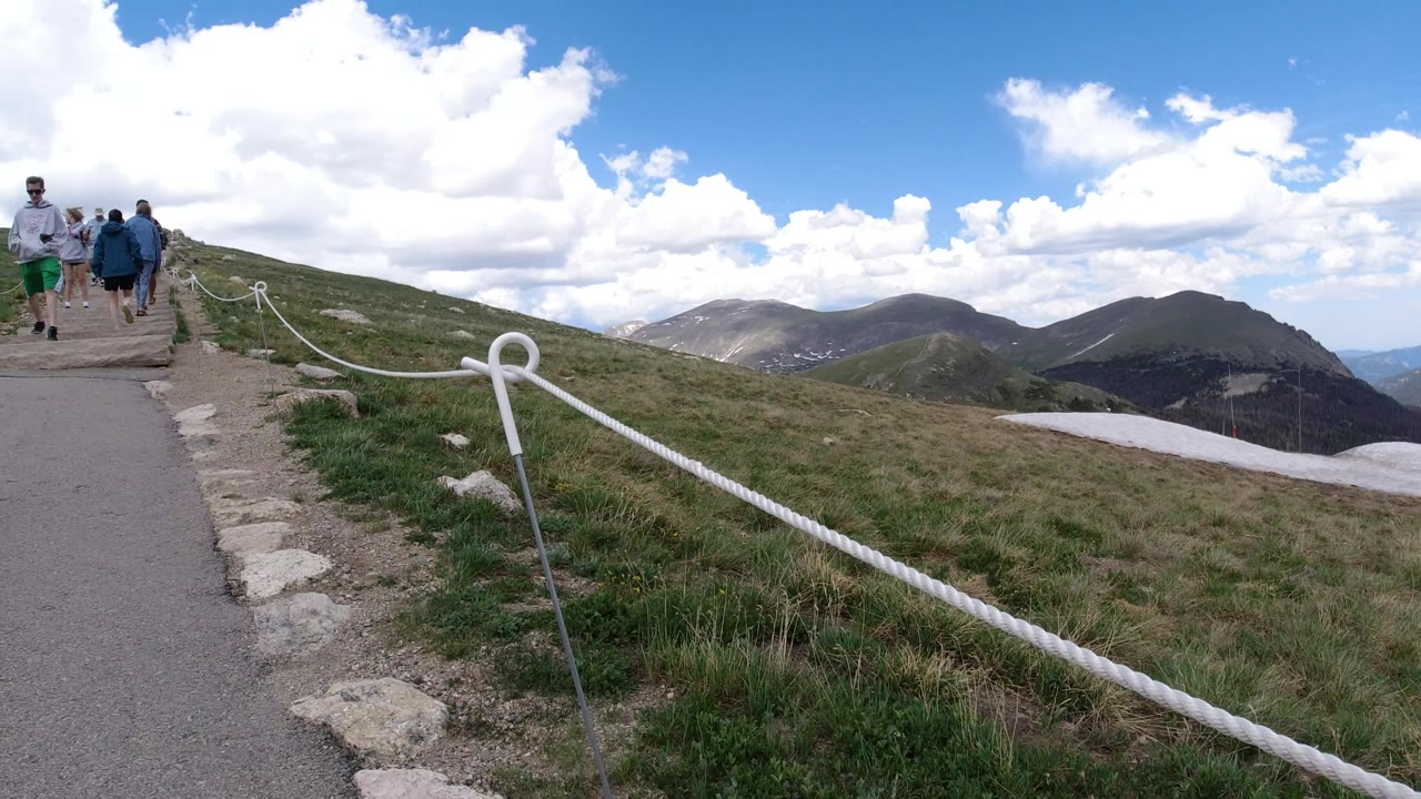 A low trail view in Winter Park, Colorado shows walkers near the left side of the path, rope railing across the foreground, grassy hillside, and rounded mountains beyond.