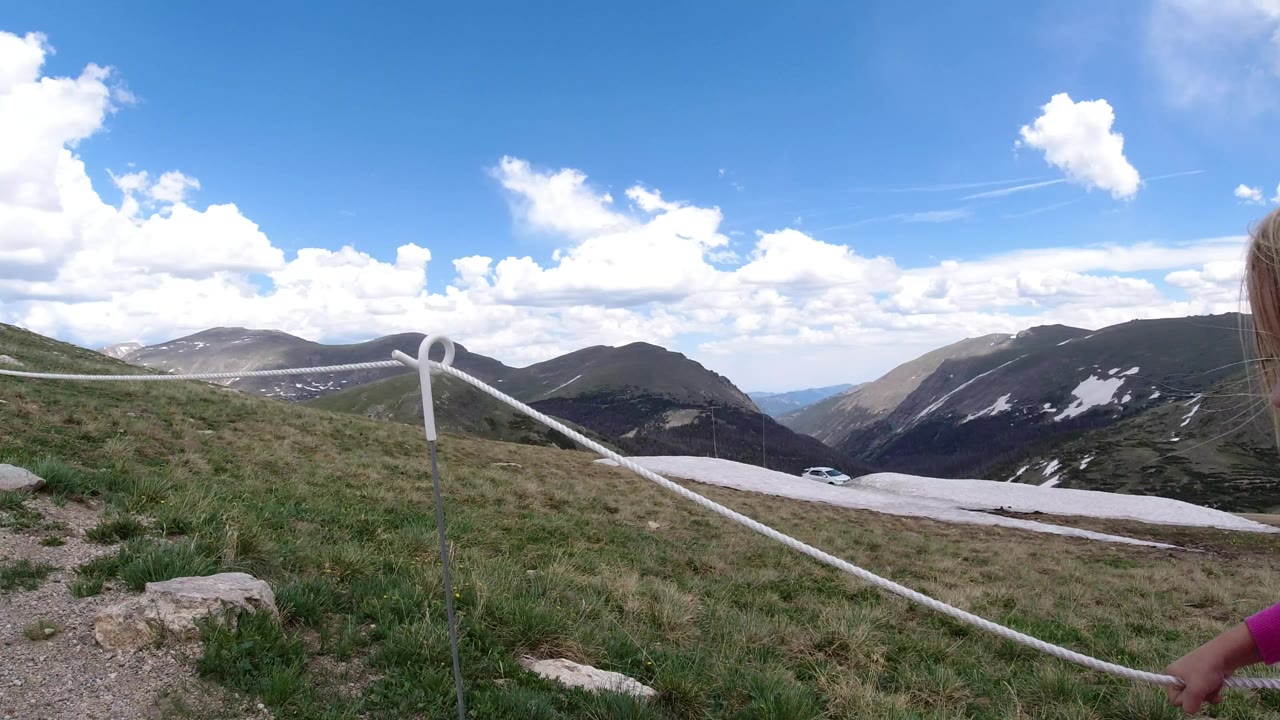 At 0:00 in Winter Park, Colorado, a roped dirt trail crosses a grassy high-country slope with distant mountain ridges, snow patches, blue sky, and no visible people.