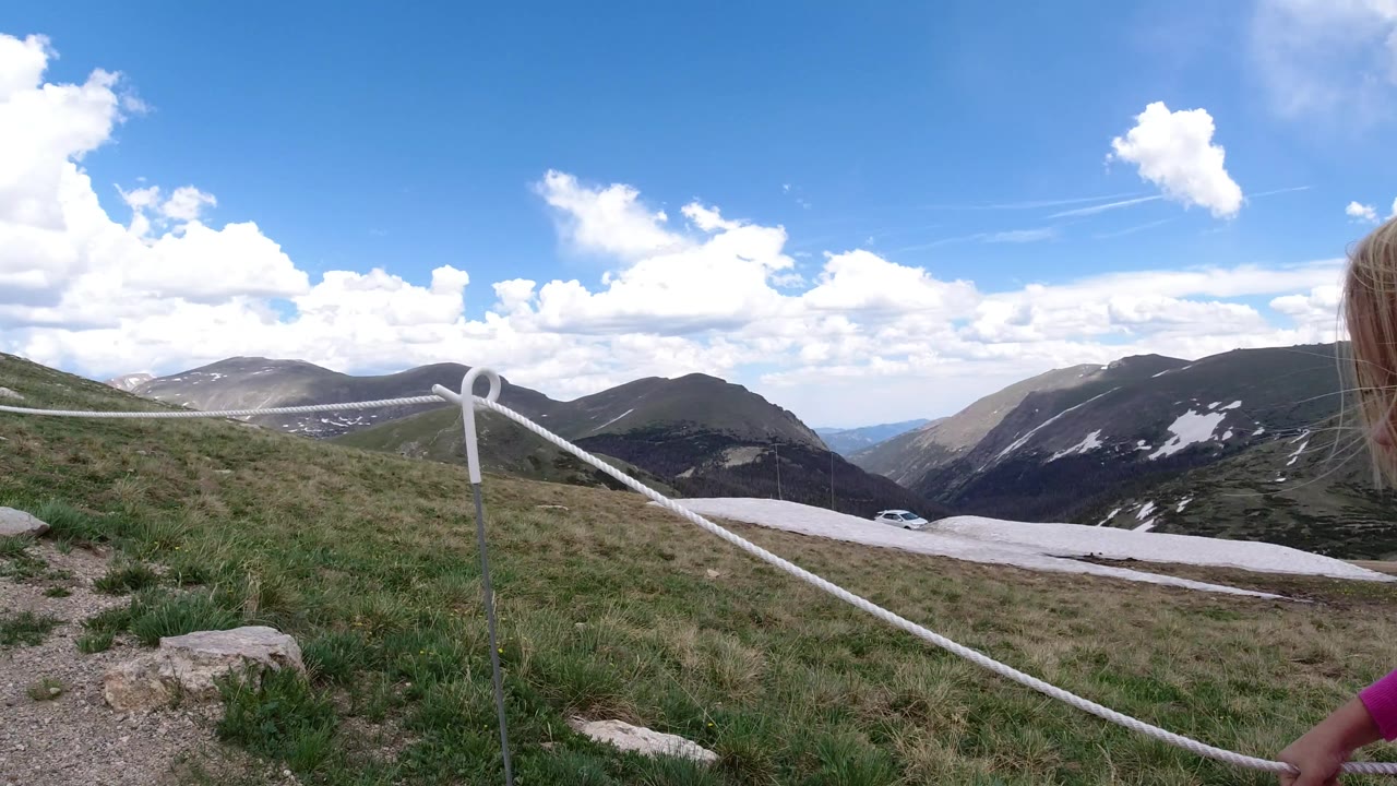 At 0:01 in Winter Park, Colorado, a child's pink sleeve and hand appear at the lower right beside the white rope barrier, with grass, rocks, snow patches, and mountains beyond.