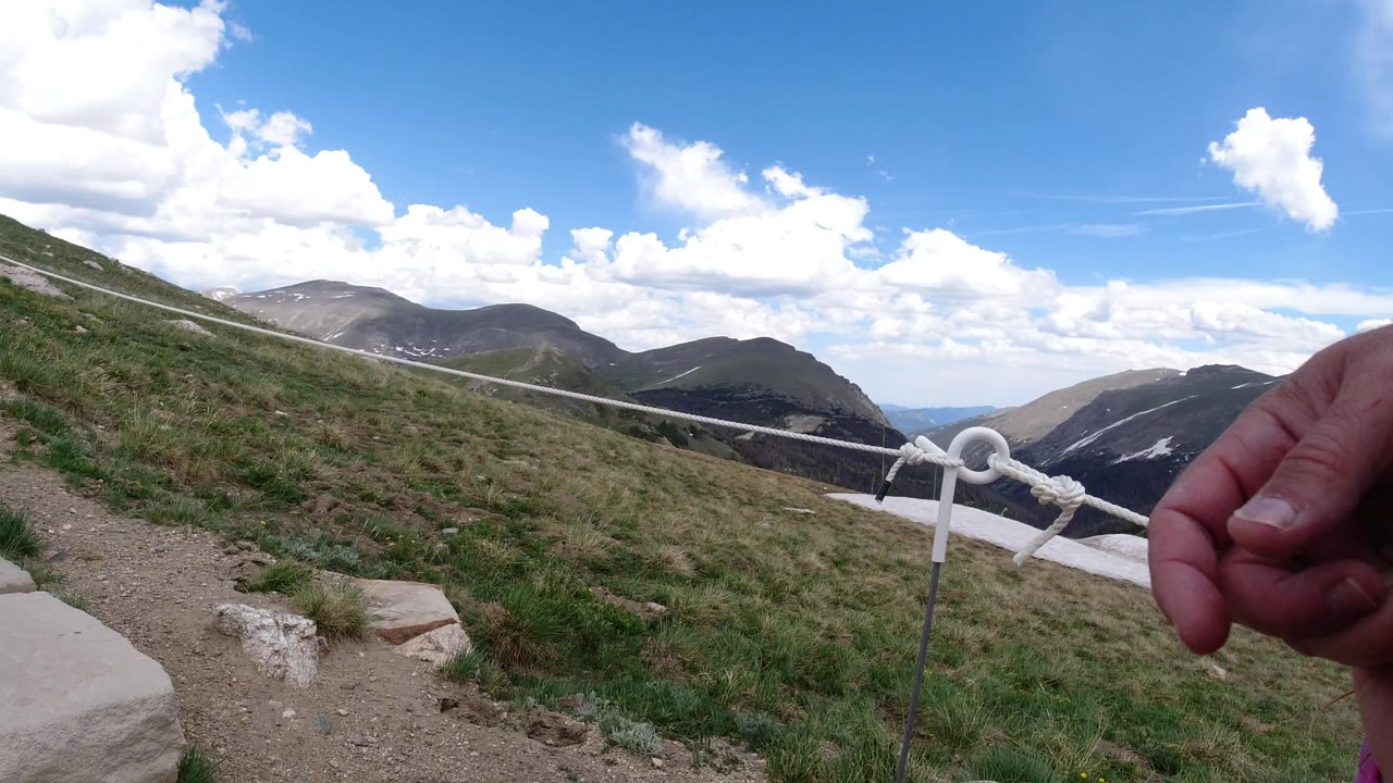 Winter Park, Colorado trail view with a white rope handrail crossing the frame, rocky steps in the foreground, mountain ridges beyond, and an adult hand partly visible at right.