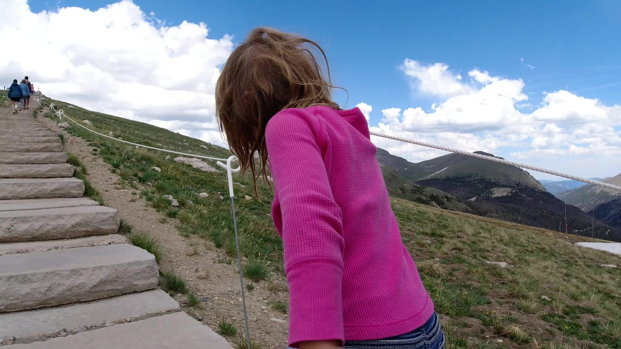 The girl remains near the right foreground in Winter Park, Colorado, looking downward beside the ascending stone steps as white rope railing cuts across the mountain view.