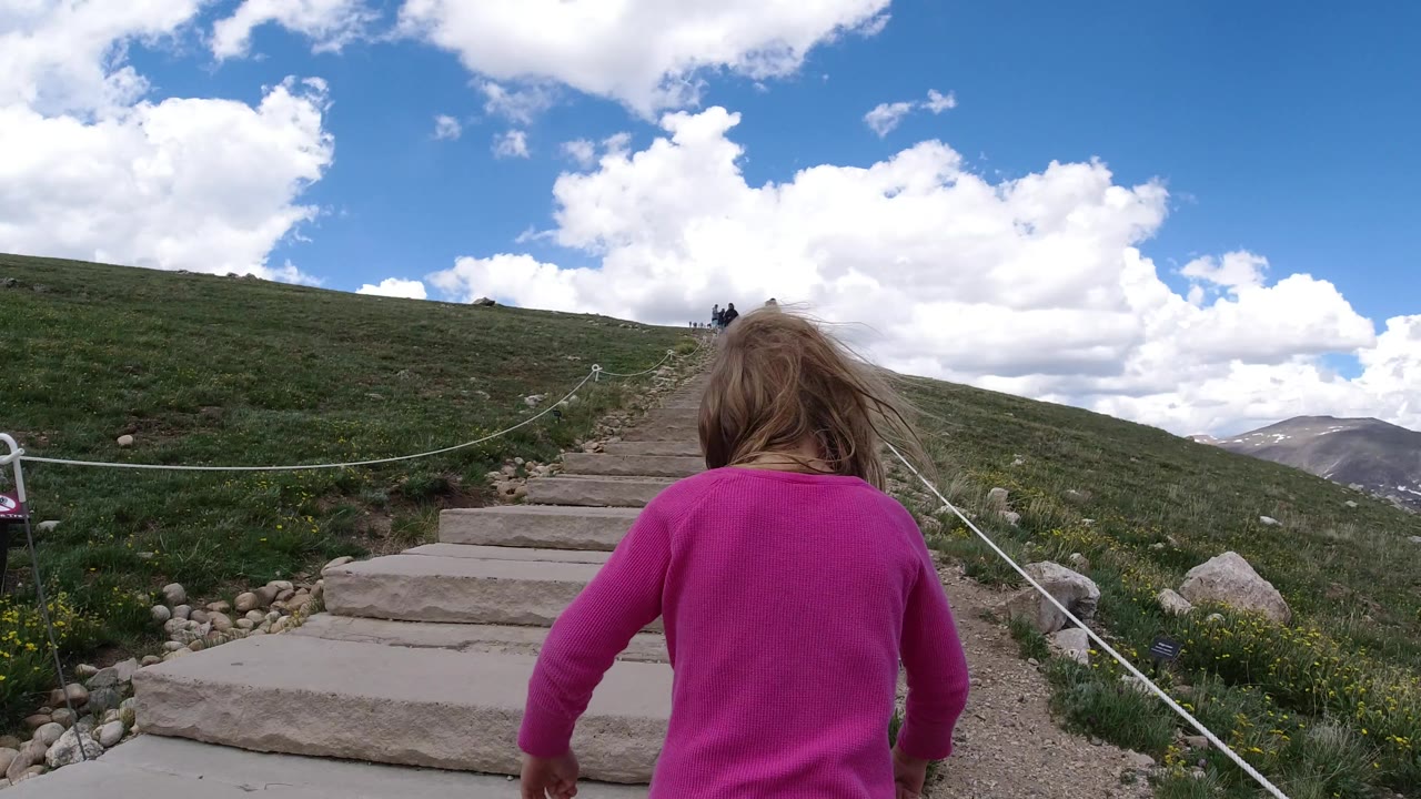 The child remains centered from behind while climbing the Winter Park, Colorado trail steps, with rope rails leading up the grassy slope and distant peaks visible to the right.