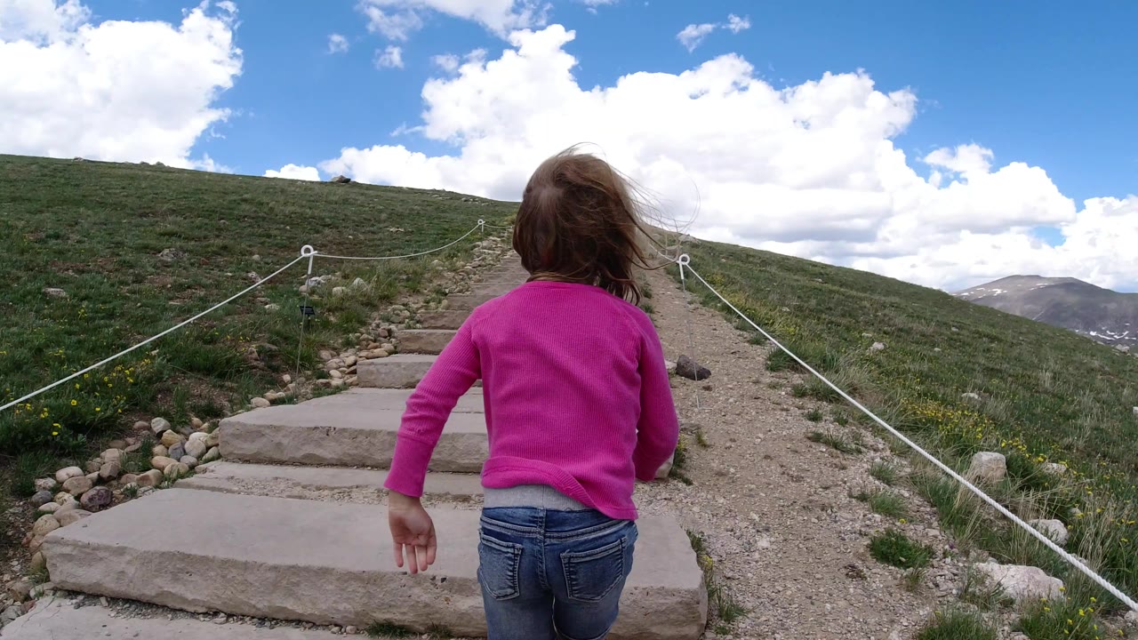 The girl walks toward the first stone steps, with white rope railings guiding the gravel path across the open Winter Park hillside.