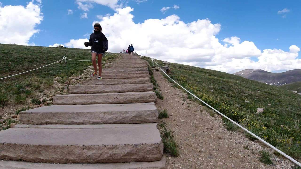 A person moves lower on the stone stairway in Winter Park, Colorado, while small hikers remain near the top and mountain ridges sit beyond the grassy slope.