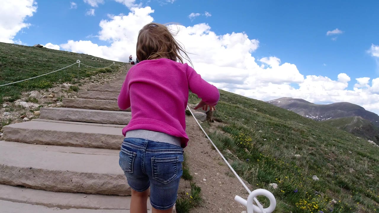 The girl remains centered from behind as she climbs the stone stair path in Winter Park, Colorado, with white rope rails leading toward people farther uphill.