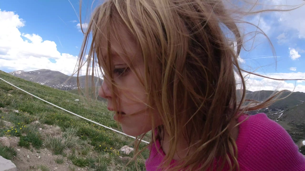 The child remains close to the lens in Winter Park, Colorado, with hair blowing across her face and a rope rail crossing the grassy hillside behind her.
