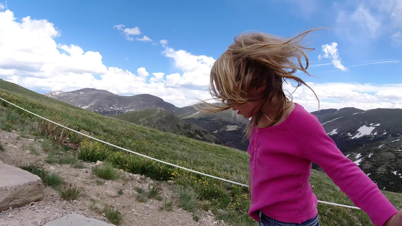 A young girl in a pink shirt stands close to the camera on stone steps in Winter Park, Colorado, with wind-blown hair, grassy slopes, distant mountains, and blue sky behind her.