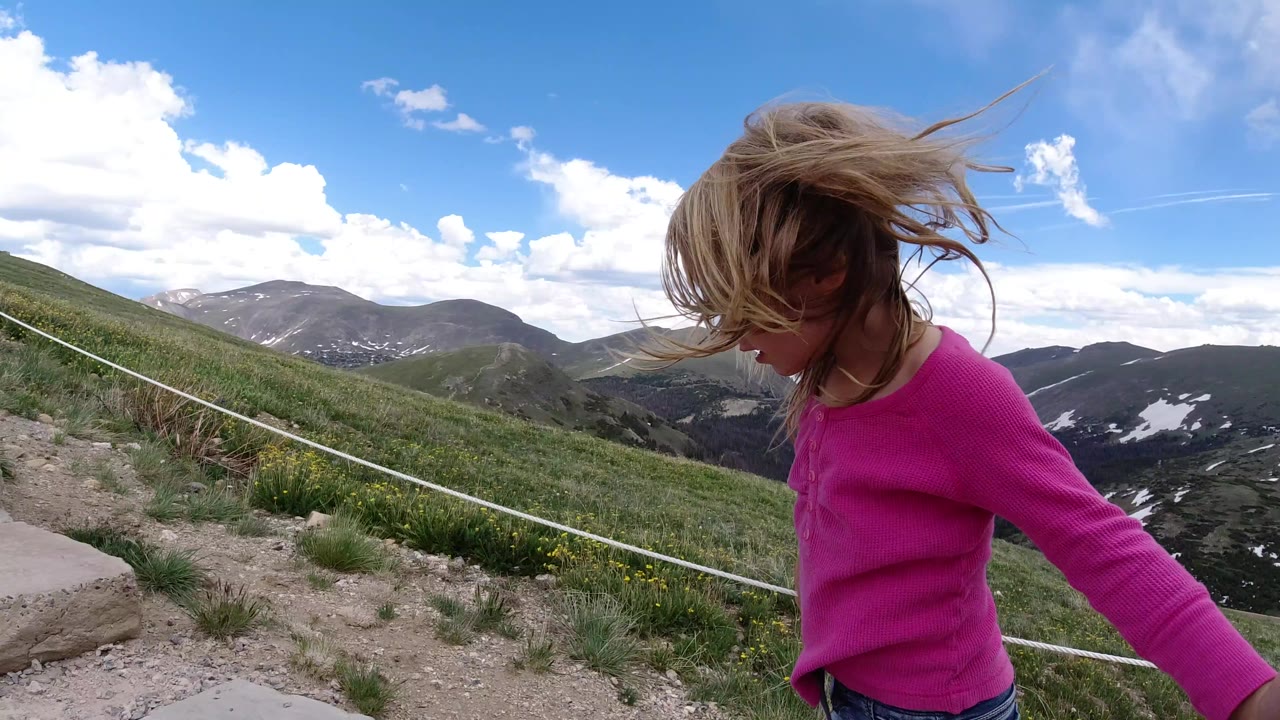 The girl remains close to the lens in Winter Park, Colorado, with her hair blown across her face while stone steps, a rope barrier, green hills, and clouded sky fill the background.