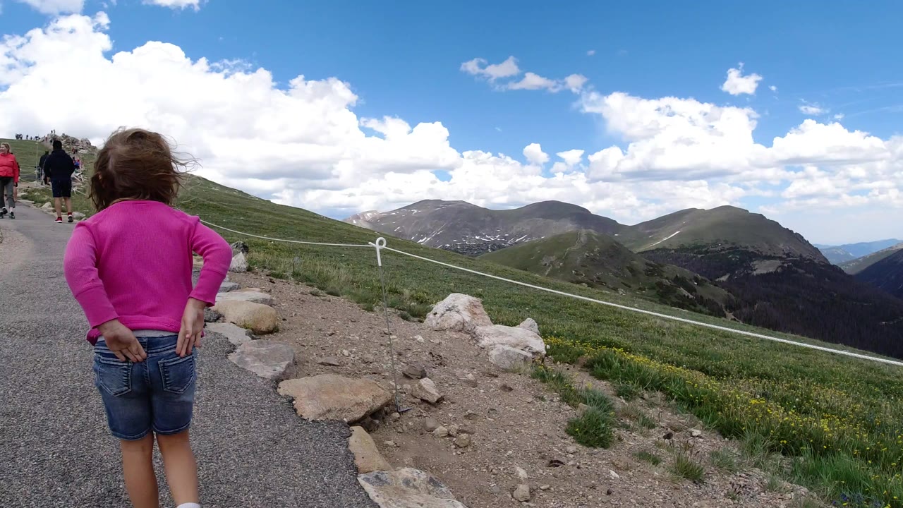 Girl in pink walking on a mountain trail with distant peaks.