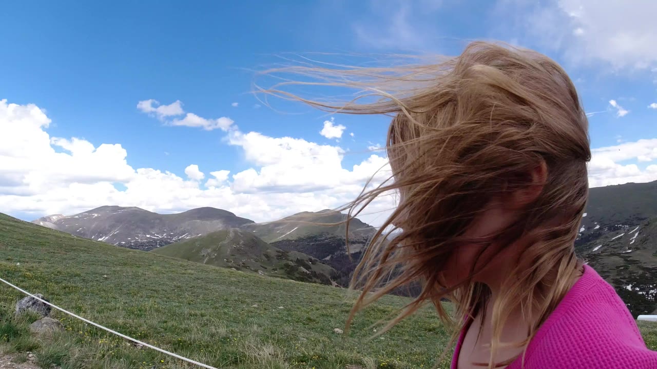 Girl in pink with windblown hair beside a mountain view.