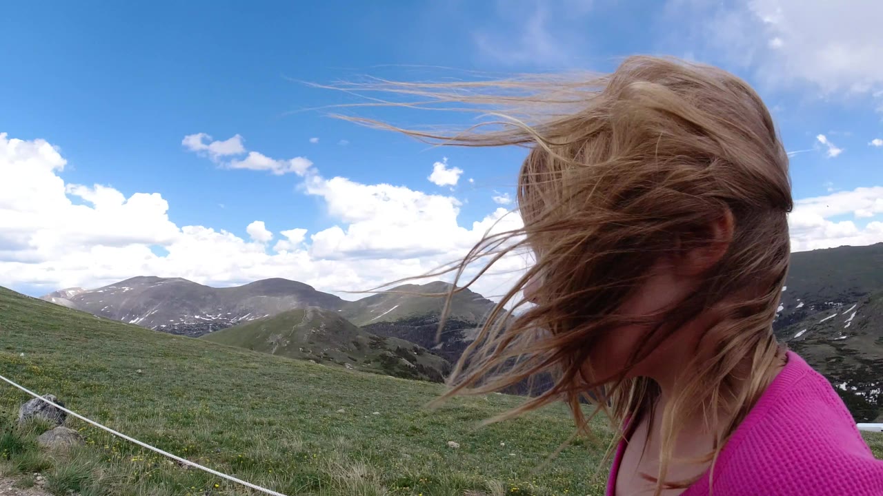 At 0:01 in Winter Park, Colorado, the girl remains near the right foreground with hair swept sideways by wind, while the grassy ridge and clouded blue sky fill the background.