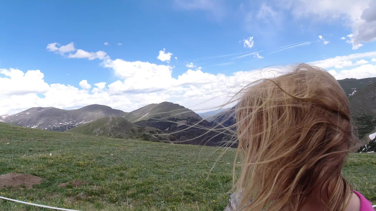 Girl with windblown blond hair facing mountain meadow.