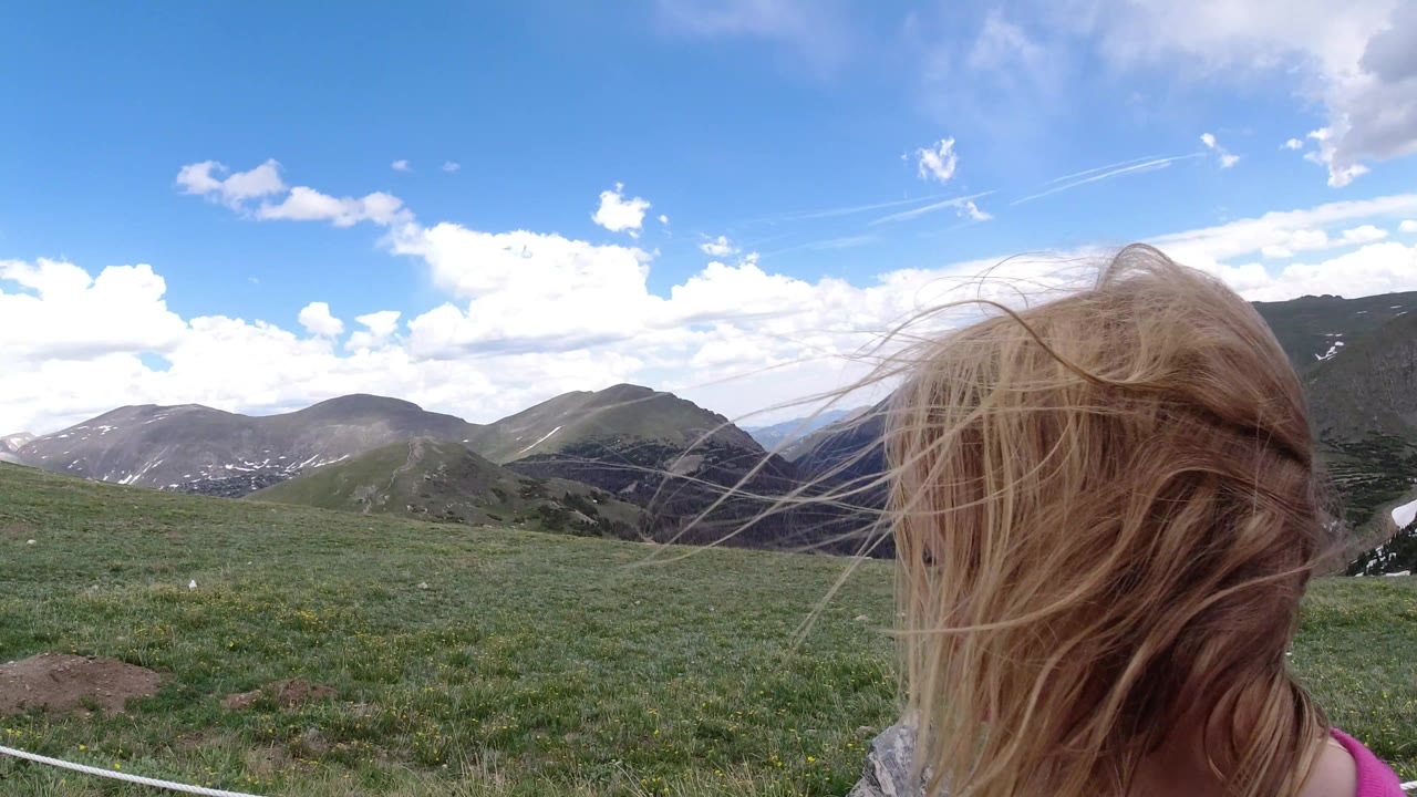 The child remains centered from behind in Winter Park, Colorado, as strands of hair cross the frame over a green meadow, low rope barrier, layered mountains, and cloud-filled sky.
