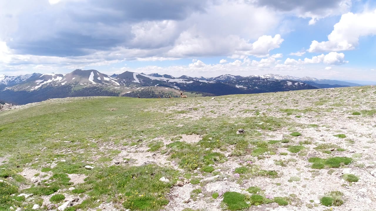 Opening frame of a Winter Park, Colorado hilltop view with pale rocky grass in front, snow-streaked mountain ridges on the horizon, large clouds overhead, and no visible people.