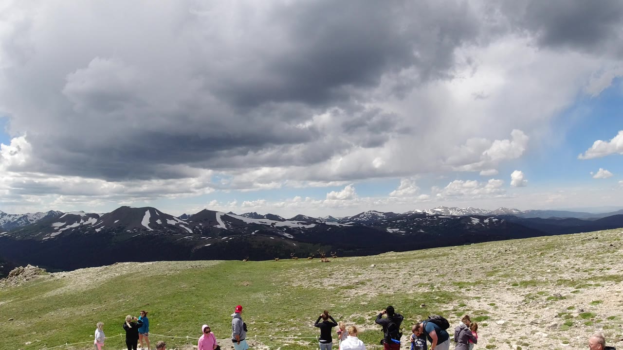 People on a grassy hilltop below dark clouds and distant snow-patched mountains.