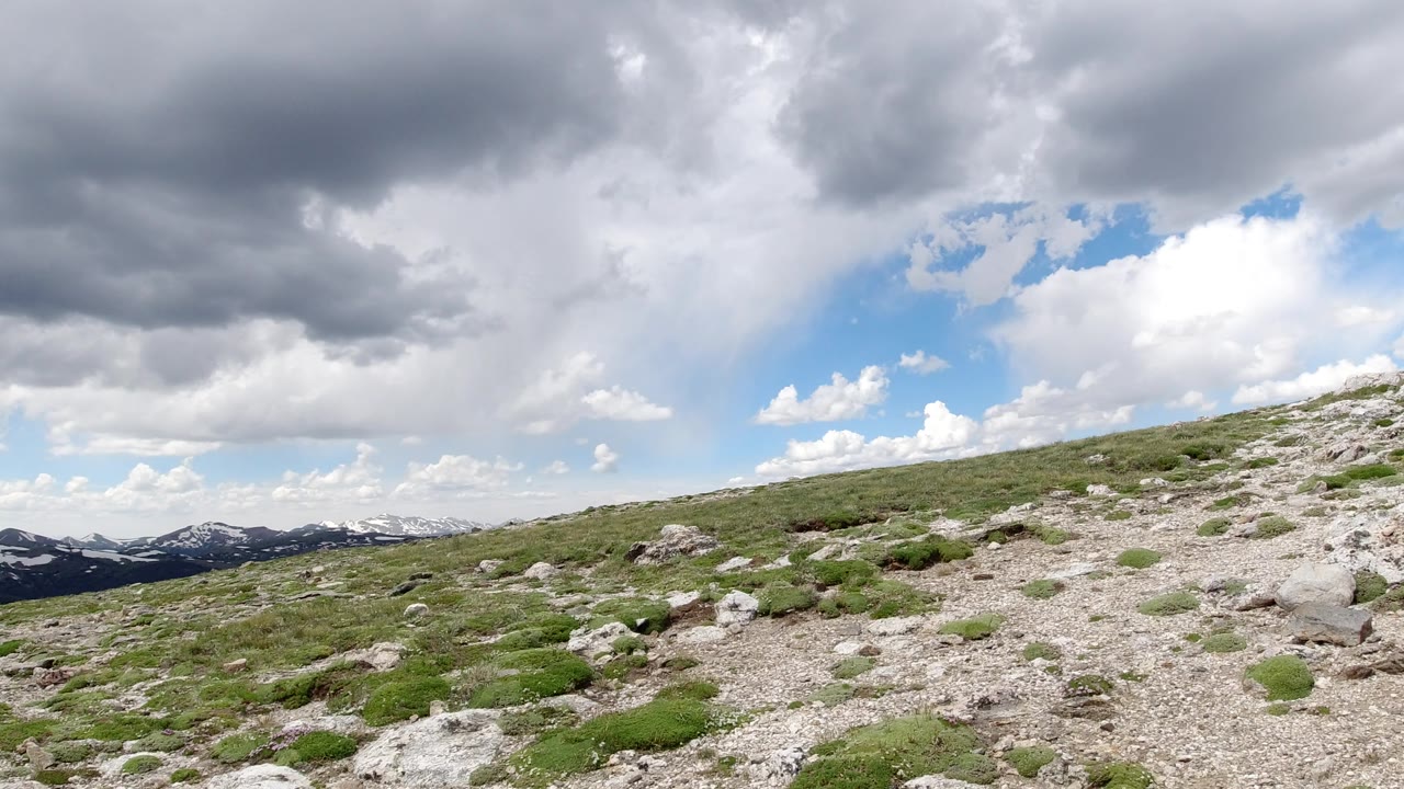 Wide view of Winter Park, Colorado rocky ground and scattered shrubs with distant snow-marked mountains low on the horizon and layered gray clouds above, with no visible people.