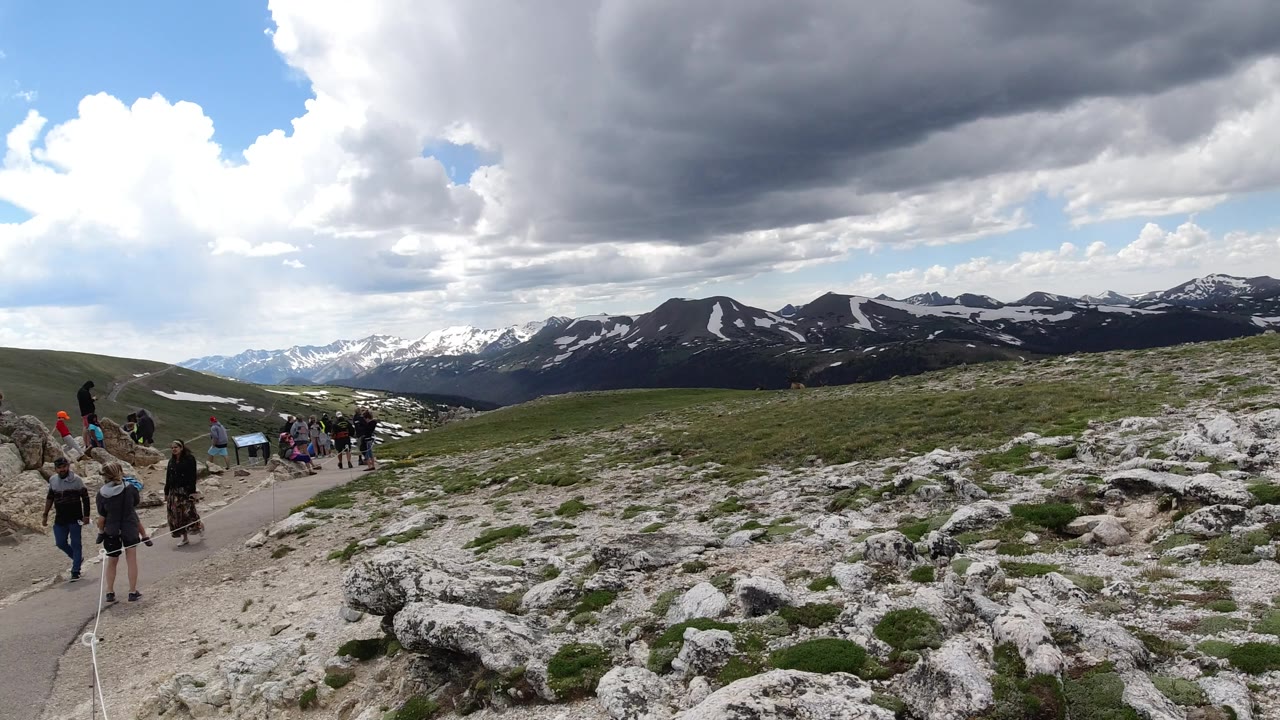 Wide Winter Park, Colorado mountain view with rocky foreground, small groups of visitors along the left trail, distant snow-streaked ridges, and a large dark cloud overhead.