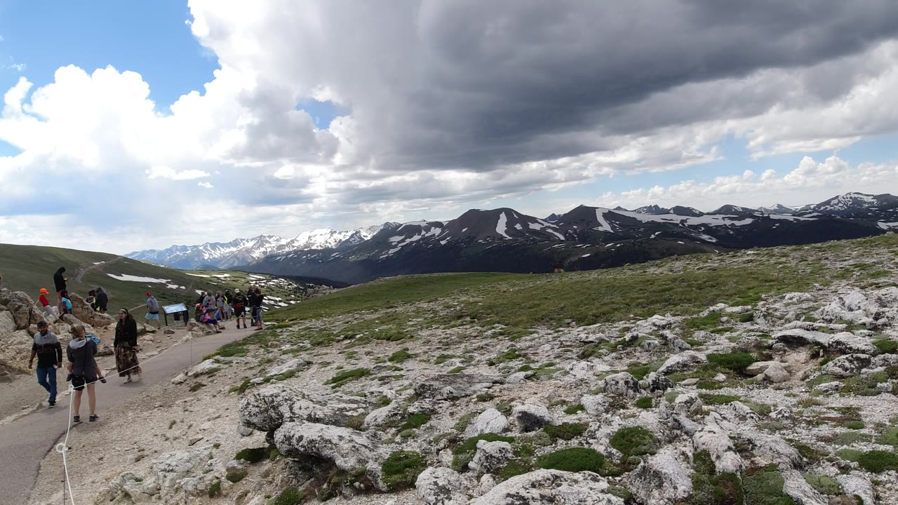 Winter Park, Colorado trail scene with several visible visitors near the left and center, pale rock slabs in the foreground, green slopes, distant mountain ridges, and mixed gray and white clouds.