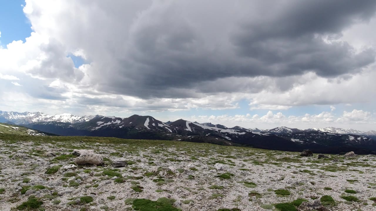 Wide Winter Park, Colorado mountain view with pale rocky ground, low green plants, distant snow-streaked ridges, heavy gray clouds, patches of blue sky, and no visible people.