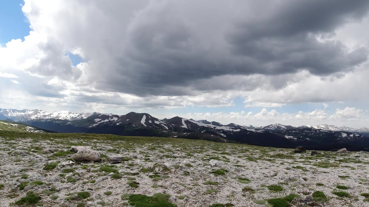 Winter Park, Colorado alpine terrain stretches across the foreground with scattered stones and green patches, while snow-marked mountains sit below a large gray cloud bank and no visible people.