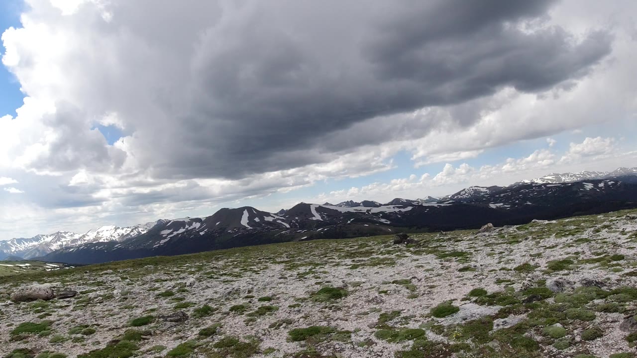Wide view in Winter Park, Colorado, with rocky grassland in front, snow-streaked mountain ridges beyond, heavy gray cloud cover overhead, and no visible people.