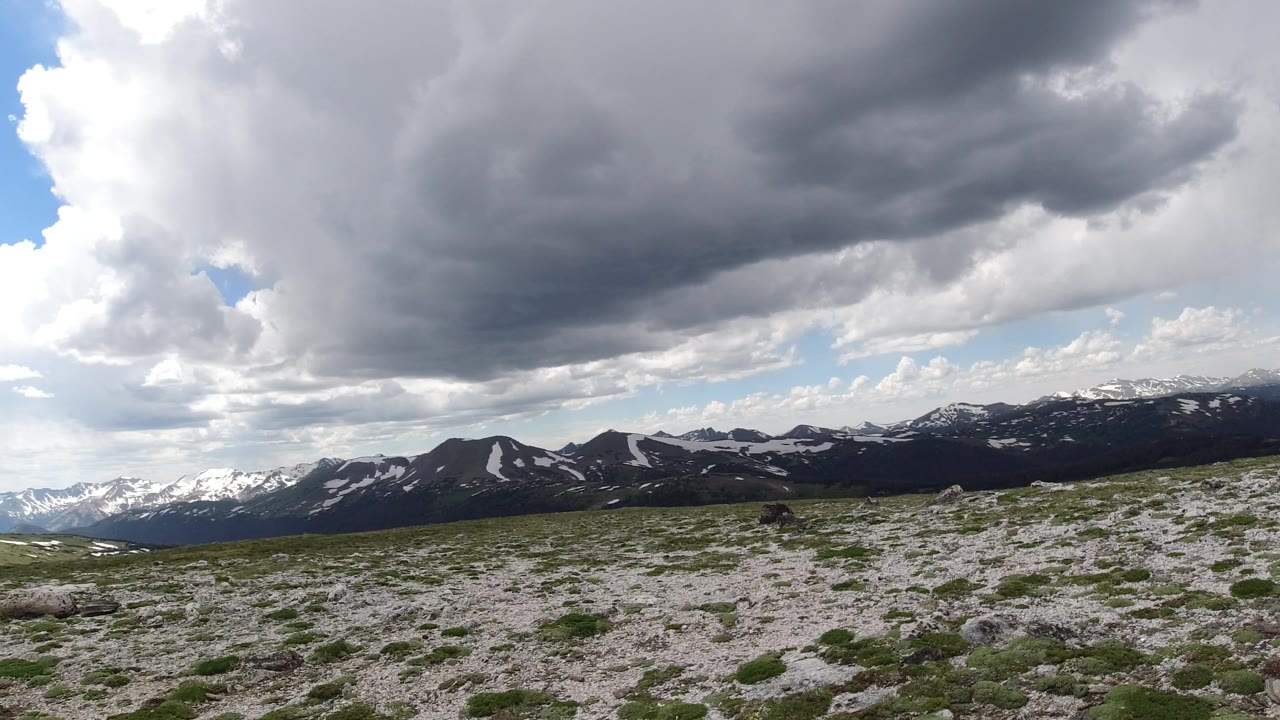 Snow-marked mountain range behind sparse grassland with stormy clouds above.