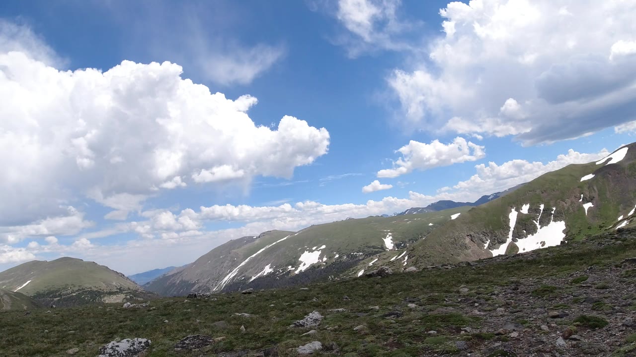 Winter Park, Colorado ridgelines remain centered behind rocky tundra, with scattered snow patches and a large white cloud mass overhead, no visible people.