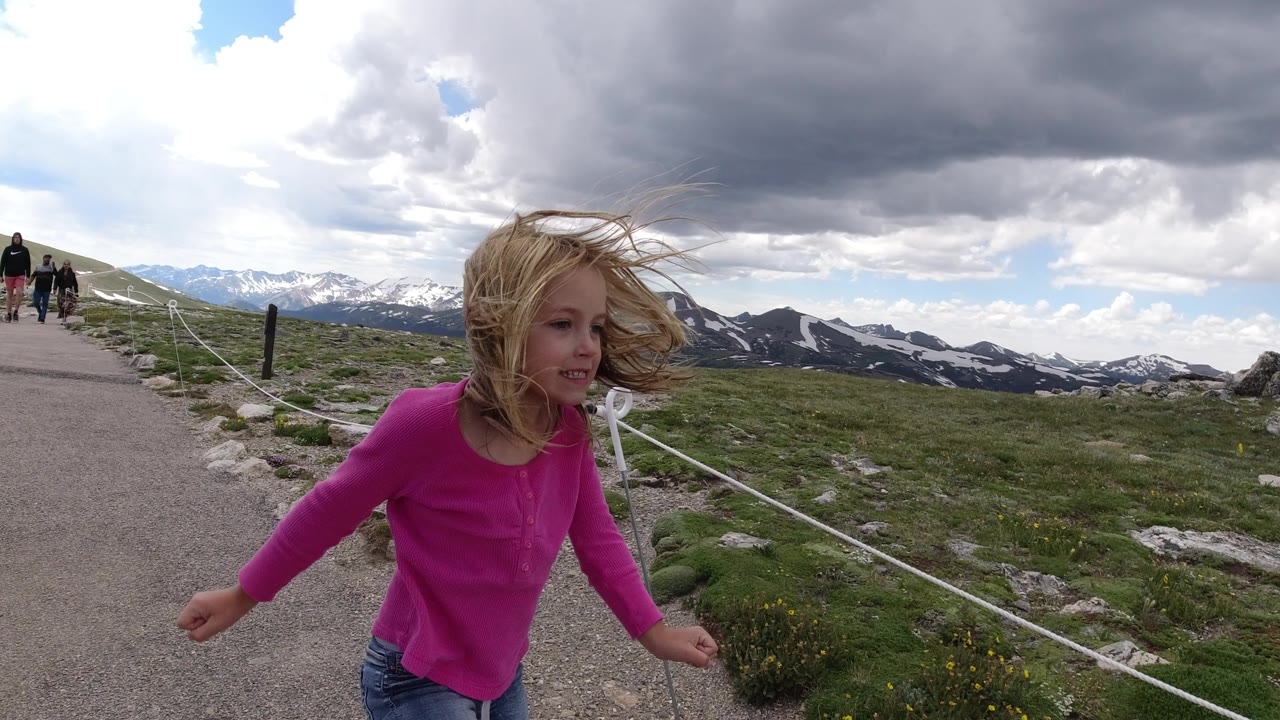 The blonde child continues along the gravel path in Winter Park, Colorado, with windblown hair, white rope fencing, green alpine vegetation, distant snow patches, and two people on the path behind her.