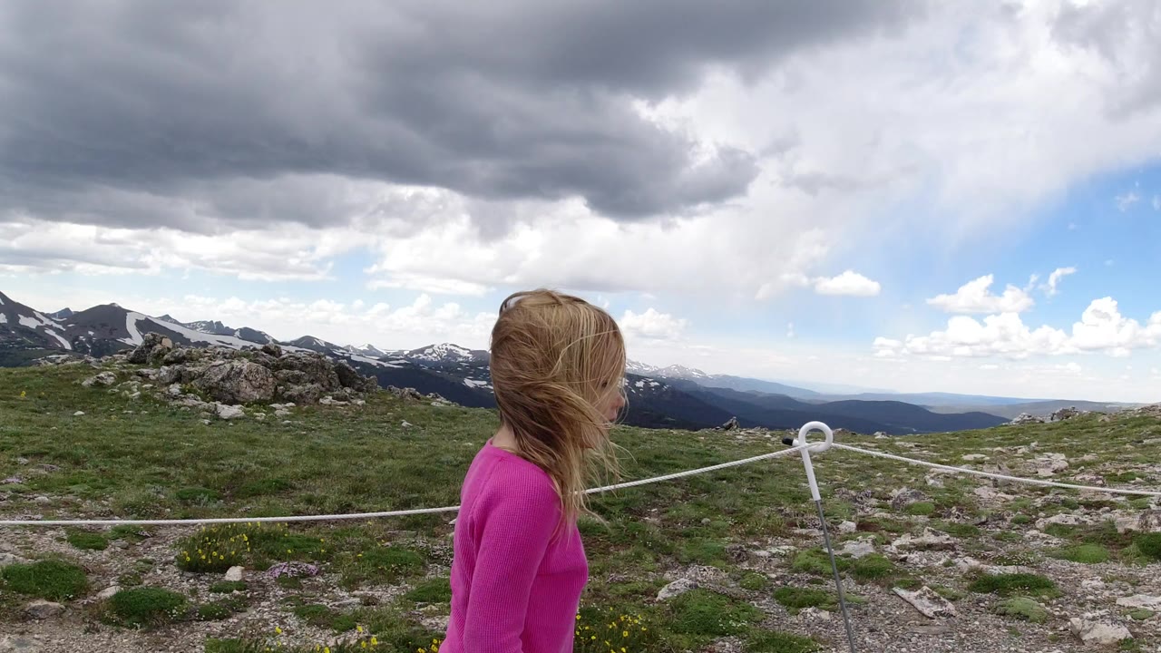 Girl in pink walking on rocky mountain trail under dark clouds.