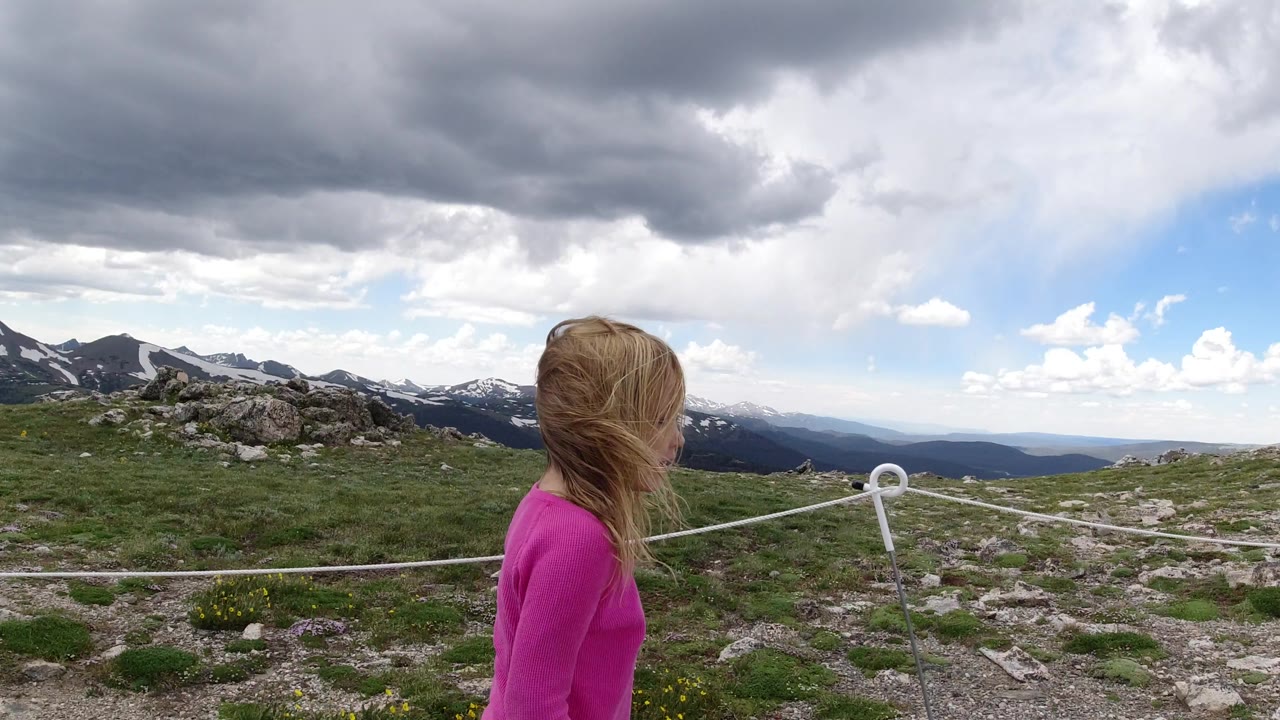 The girl is seen mostly from behind beside the white rope boundary on the Winter Park, Colorado trail, with low rocks, green ground, distant mountains, and layered storm clouds.
