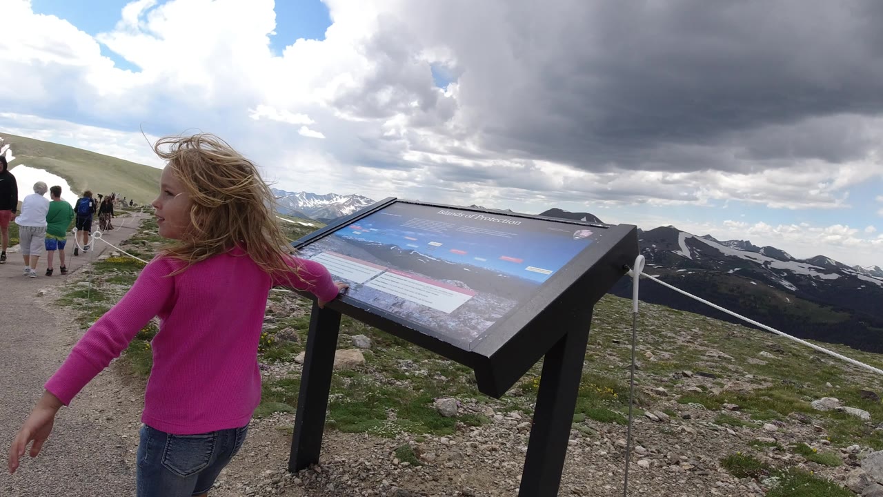 The girl turns beside the angled sign while a small tourist group continues along the Winter Park, Colorado overlook path with dark mountain ridges behind them.