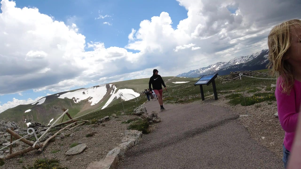 Child and adult on a mountain trail with snow patches and clouds.