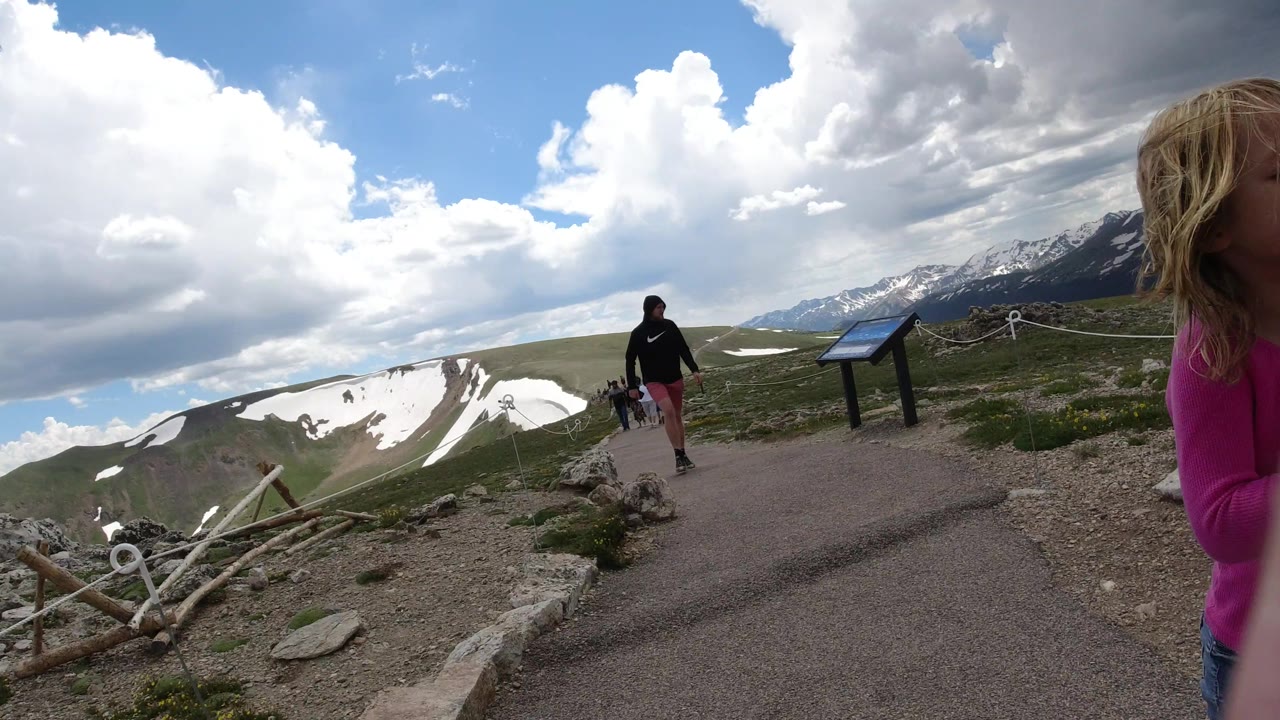The handheld view stays low and wide on a Winter Park, Colorado trail, with the child partly at frame right, an adult ahead, a sign, rope barrier, snow patches, and mountains.