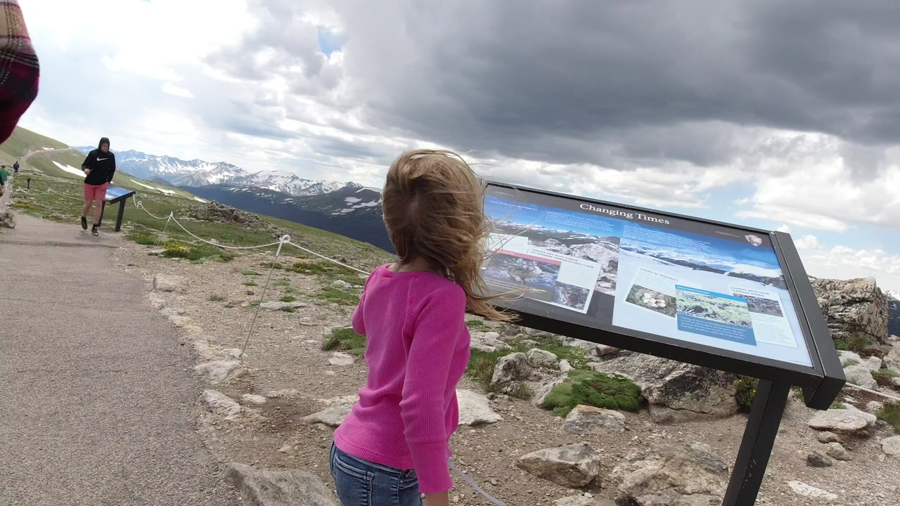 Child in pink standing near a mountain sign board under cloudy sky.