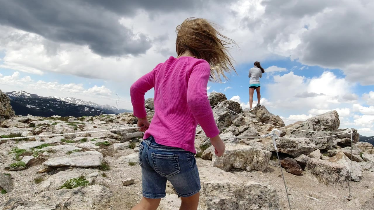 The child remains close to the camera as the rocky path, rope posts, adult figure, mountain ridge, and mixed cloud cover fill the Winter Park, Colorado frame.