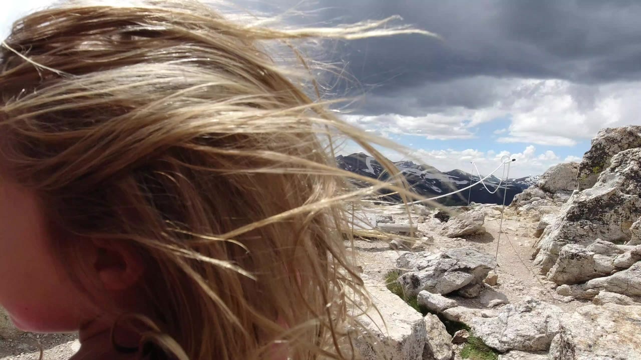 At the start of the Winter Park, Colorado clip, windblown blonde hair and the edge of a child’s face fill the left foreground beside pale rocks, a rope barrier, distant mountains, and dark clouds.