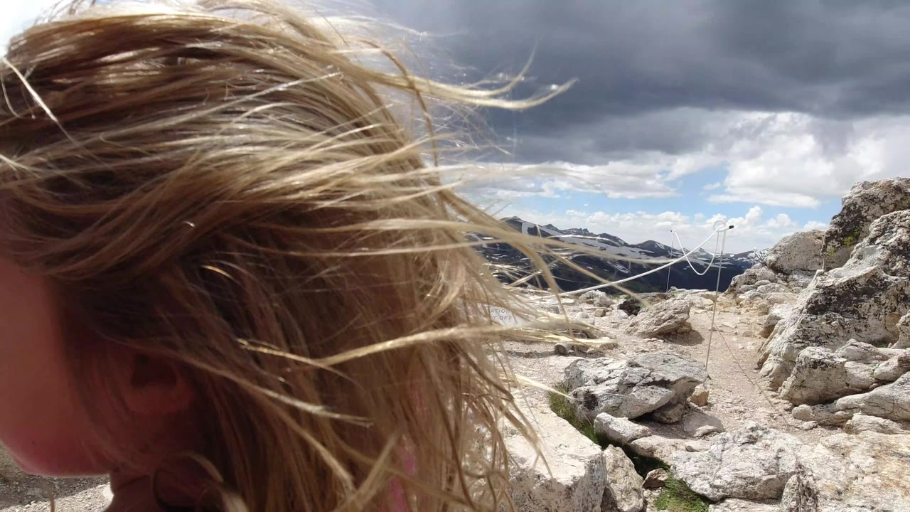 Windblown blonde hair and the side of a child’s head dominate the foreground in Winter Park, Colorado, with rough rocks, rope lines, mountain ridges, and heavy clouds beyond.